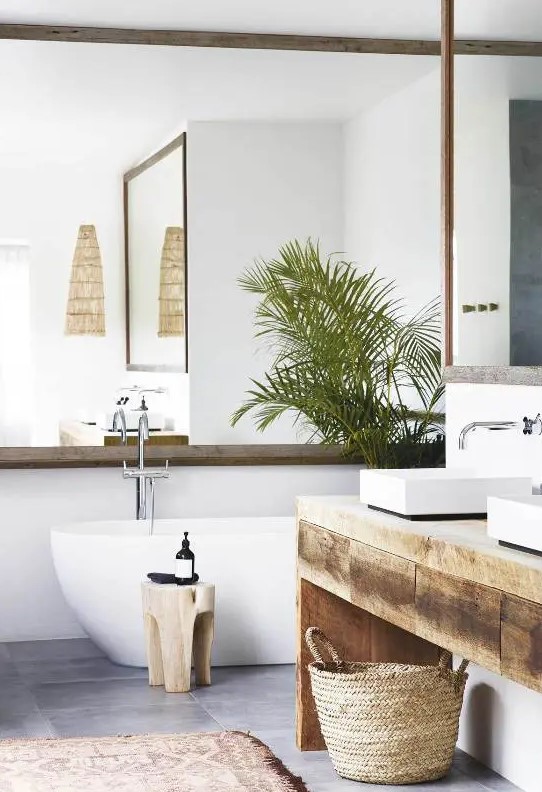 a contemporary tropical bathroom with a mirror that takes a whole wall, a wooden vanity, a tub, a basket and some potted plants