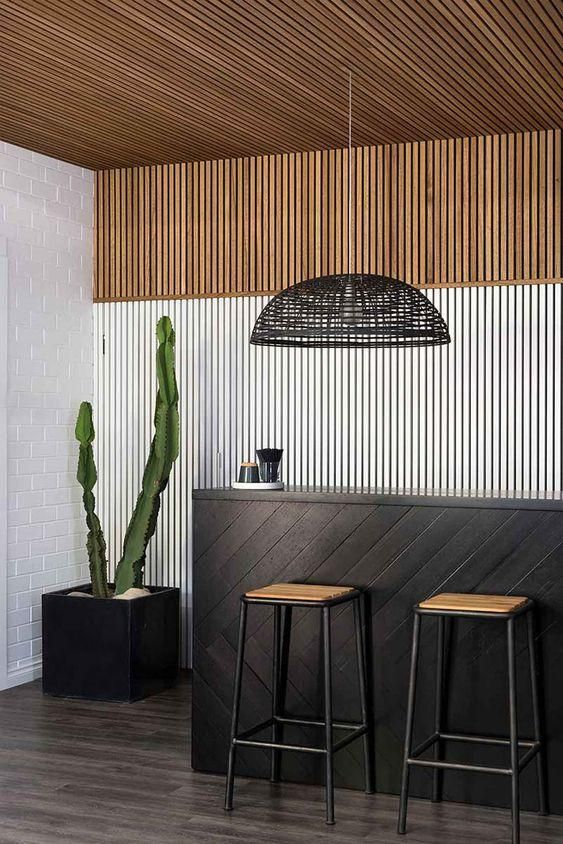 a cool home bar with a wood slat accent wall, a black bar counter, tall stools, a black pendant lamp and a potted cactus