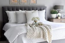 a farmhouse bedroom with grey walls, a grey upholstered bed with white bedding, a grid gallery wall of vintage herbs and blooms
