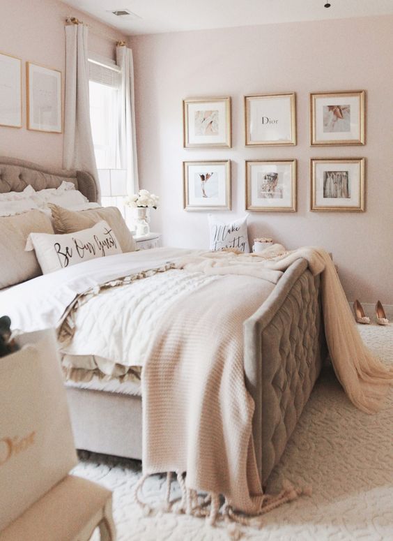 a glam bedroom with light pink walls, a grey upholstered bed with neutral bedding, a grid gallery wall and another one over the headboard