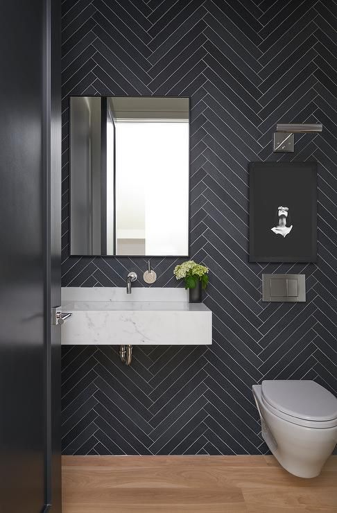 a laconic and moody bathroom clad with navy herringbone tiles, a floating sink of white stone, a rectangular mirror and an artwork is chic and cool