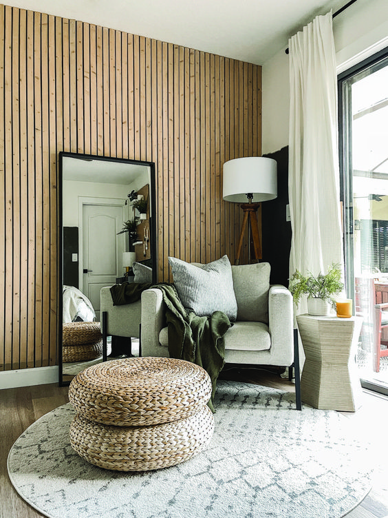 a lovely boho space with a wood slat accent wall, a neutral chair, a floor lamp, a mirror, layered rugs and jute poufs