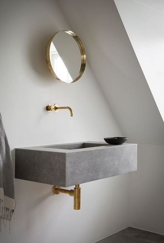 a minimalist attic bathroom featuring a floating grey stone sink, brass fixtures and a round mirror plus lots of natural light