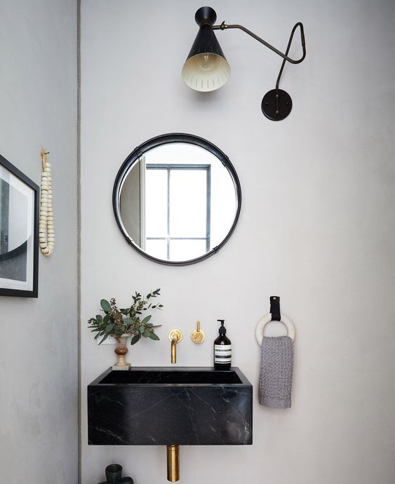 a modern and catchy bathroom with a black floating sink, a round mirror, a black retro sconce and some greenery in a vase