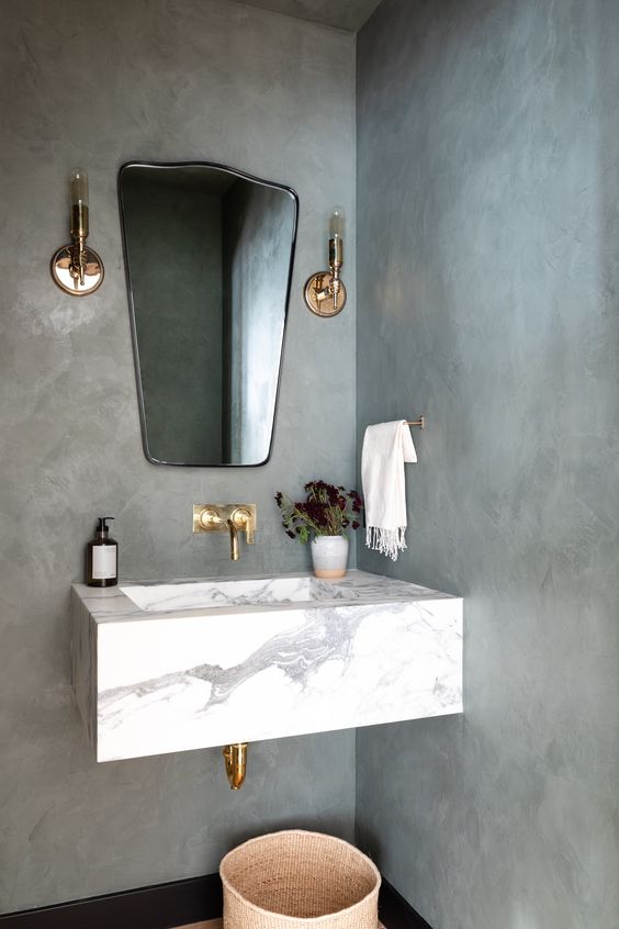 a modern bathroom with cocnrete walls, a small yet chic marble slab floating sink, brass fixtures and a catchy mirror