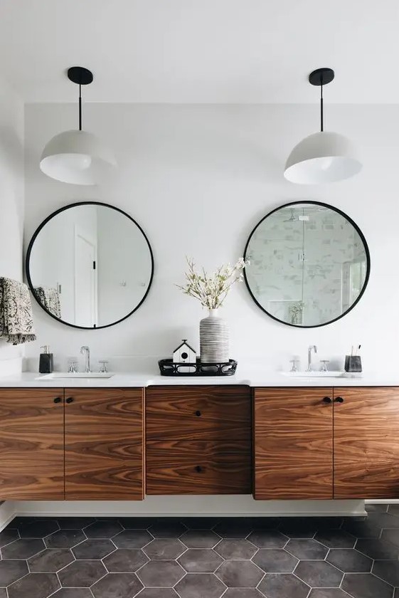 a modern bathroom with grey hex tiles on the floor, a rich-toned floating timber vanity and round mirrors