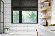a modern country bathroom with white subway tiles and black herringbone ones, a bathtub clad with white marble, light stained furniture and a window with a shade