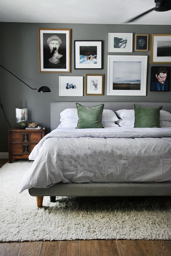 a moody grey bedroom with a grey upholstered bed with grey and grene bedding, stained nightstands, a free form gallery wall