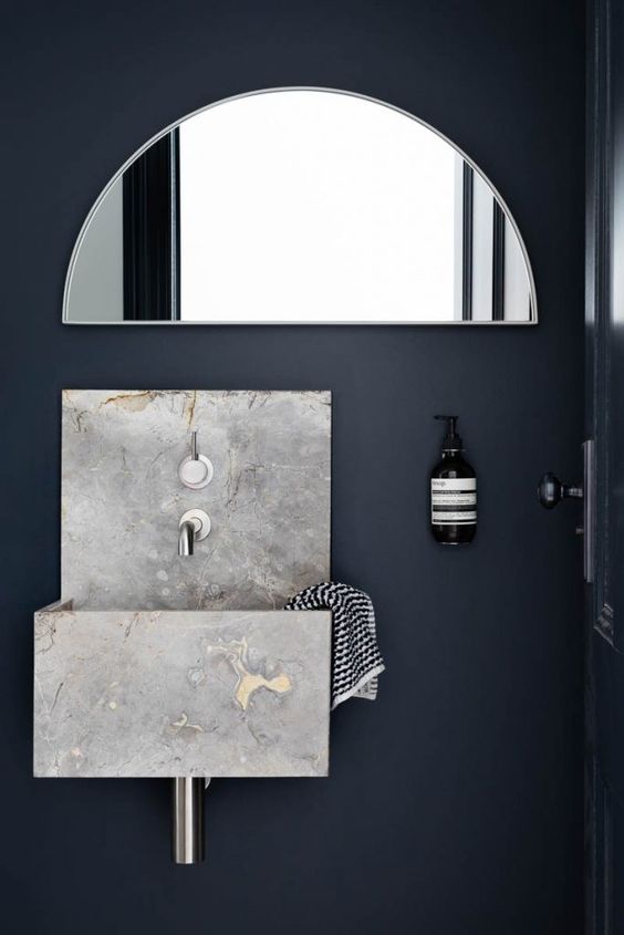 a navy powder room with a grey stone floating sink, a semi circle mirror, a soap bottle attached to the wall