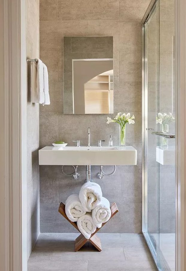 a neutral bathroom with a floating sink, a stand with towels, a mirror with no frame and a shower space enclosed in glass