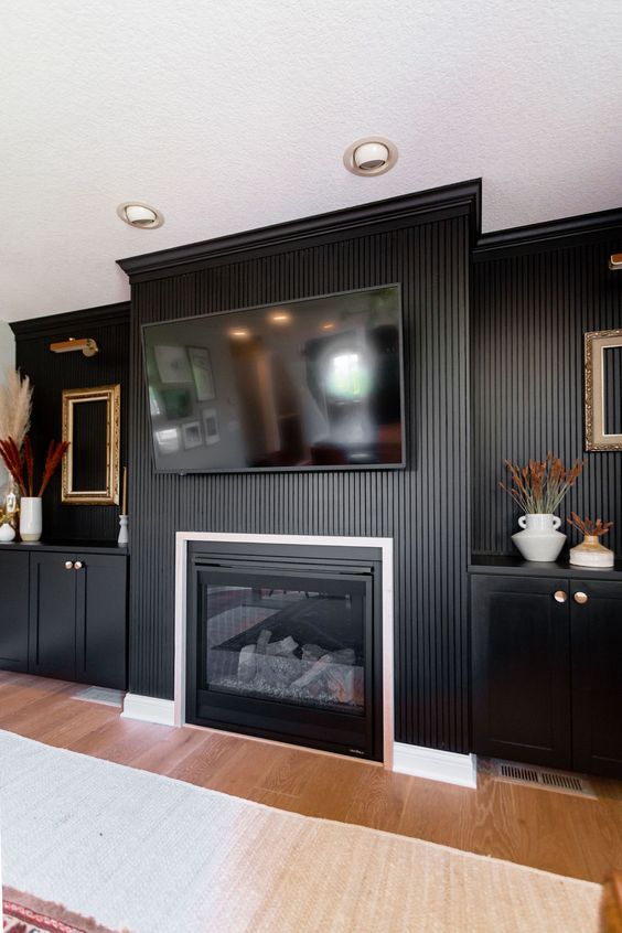 a refined living room with black walls and a wood slat accent one, a built-in fireplace and black cabinets, gold touches