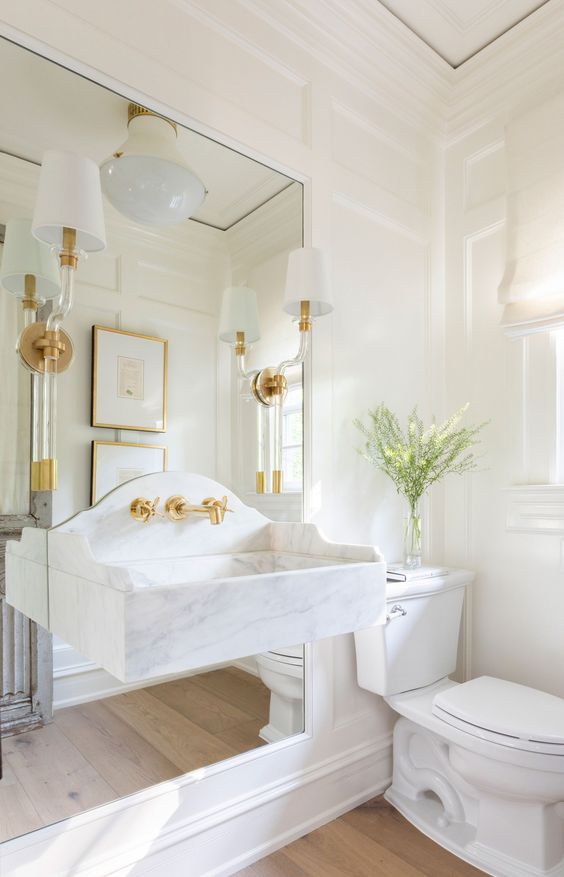 a refined white bathroom with paneled walls, a mirror and a floating stone sink attached to it, some sconces and lamps and white appliances