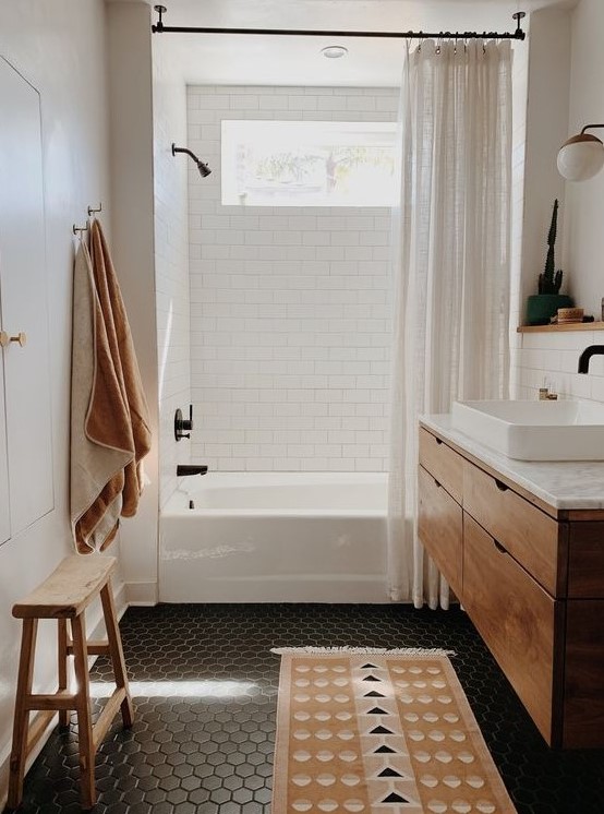 a small and cozy bathroom done with white subway and black hex tiles, a floating timber vanity, neutral linens and a wooden stool