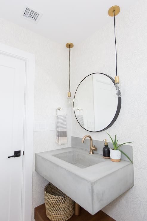 a small powder room with a concrete sink and vanity in one, a round mirror, pendant bulbs, a basket and brass and gold fixtures