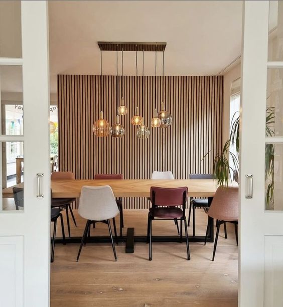 a stylish dining room with a wood slat wall, a stained table, mismatching chairs, a cluster of pendant lamps and potted plants