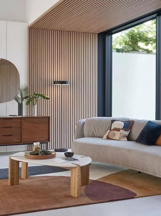 an elegant mid-century modern living room with a wood slat accent wall and ceiling, a curved sofa, a printed rug, a stained credenza and a coffee tabl