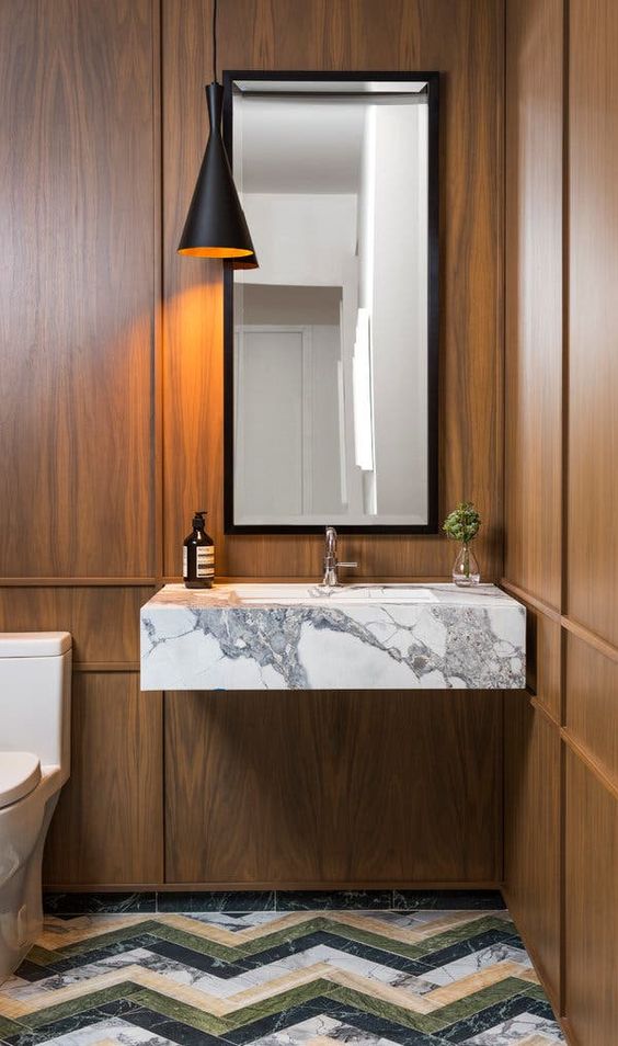 an elegant powder room with wood panels on the walls, a floating white marble sink, a catchy chevron marble floor and a black sconce