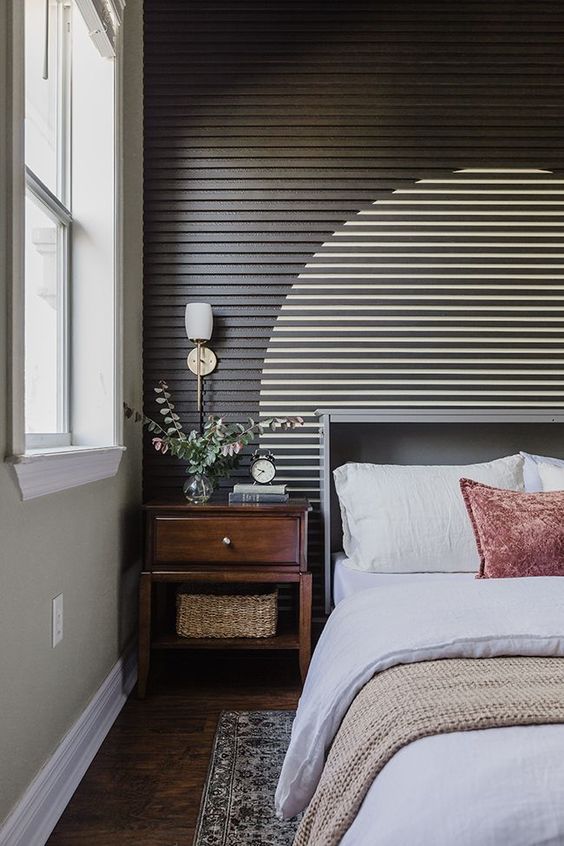 an eye-catching wood slat accent wall with a painted circle, a grey bed with bedding, a stained nightstand and some printed rugs