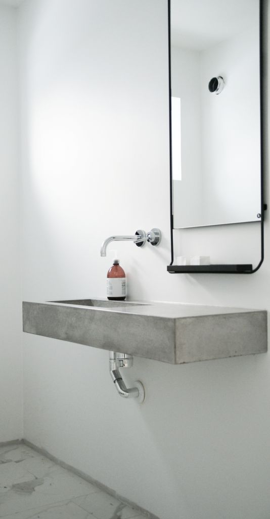 an ultra-minimalist bathroom with white walls, a concrete floating sink, a catchy mirror with an additional small shelf