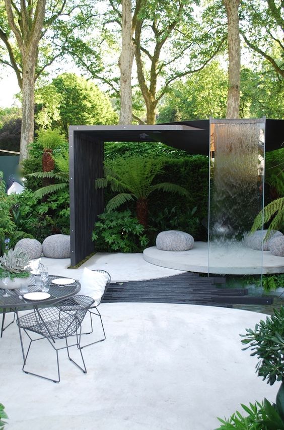 a contemporary outdoor space with a concrete round deck, a dining space, a round platform with pebbles and a waterfall of glass