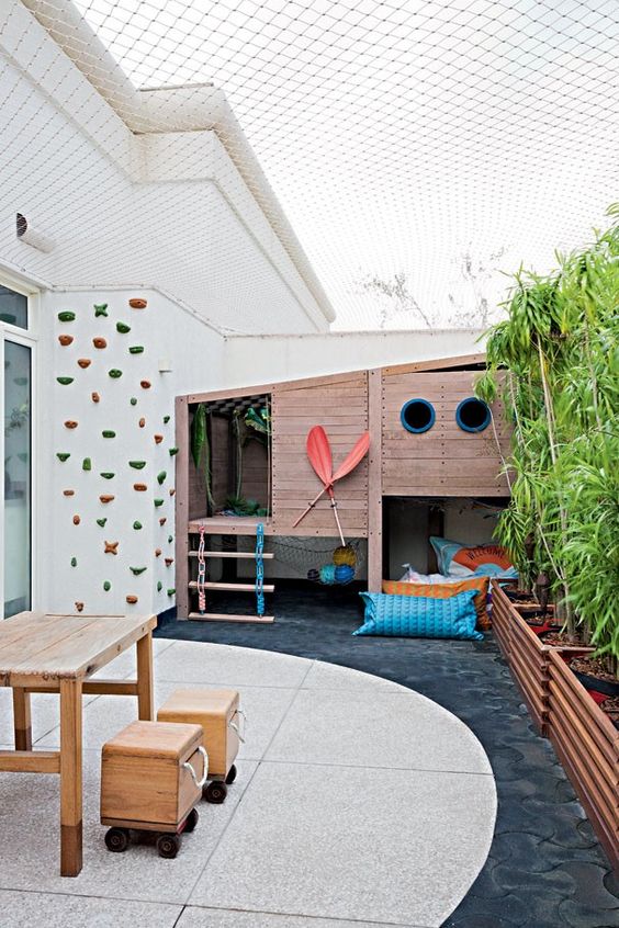 a kids' backyard with a house, a lounge zone with lots of pillows and a climbing wall, a dining set of wood