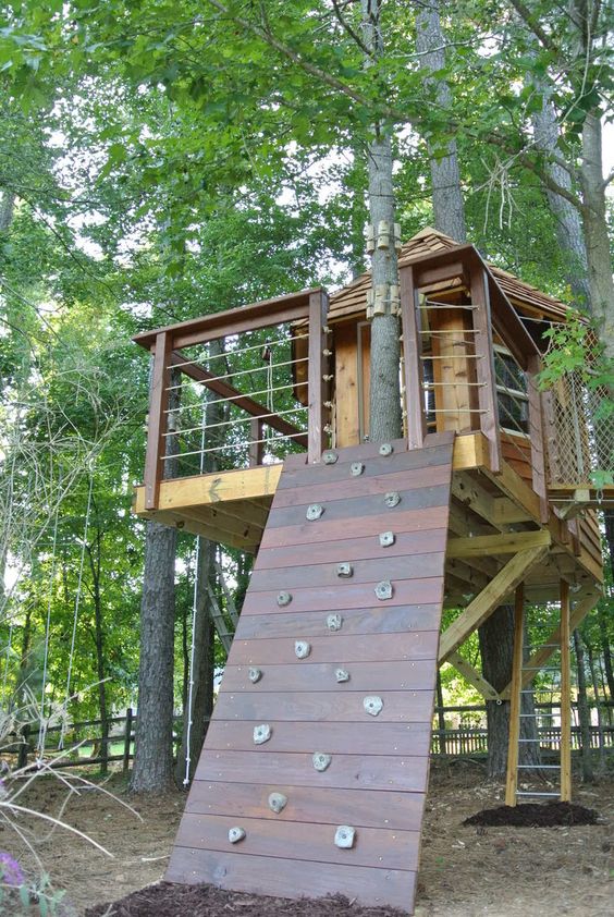 a gorgeous stained wood tree house with living trees, a climbing wall and a ladder is a lovely idea for a rustic outdoor space