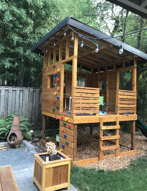 a kids' playhouse of wood, with a roof and some furniture, a colorful cimbing wall, a ladder and some planters around is a cool space a kids' playhouse of wood, with a roof and some furniture, a colorful cimbing wall, a ladder and some planters around is a cool space