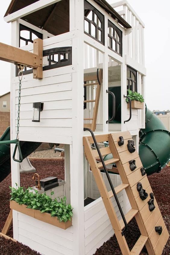 a white farmhouse playhouse for kids, with ladders, potted greenery and a climbing wall for a bit of fun