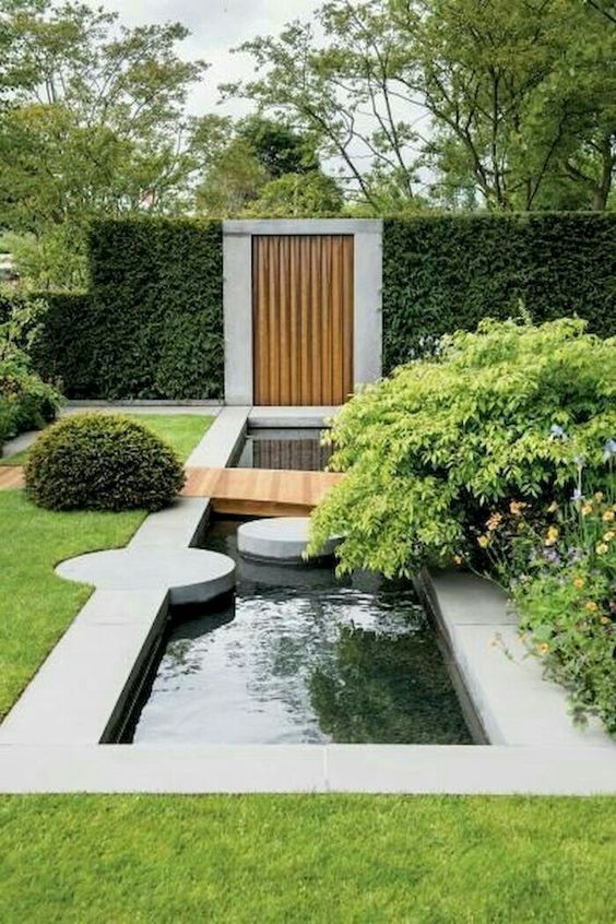 a modern garden with lots of greenery and bushes, with a water feature, stone pavements and a pathway over the water feature