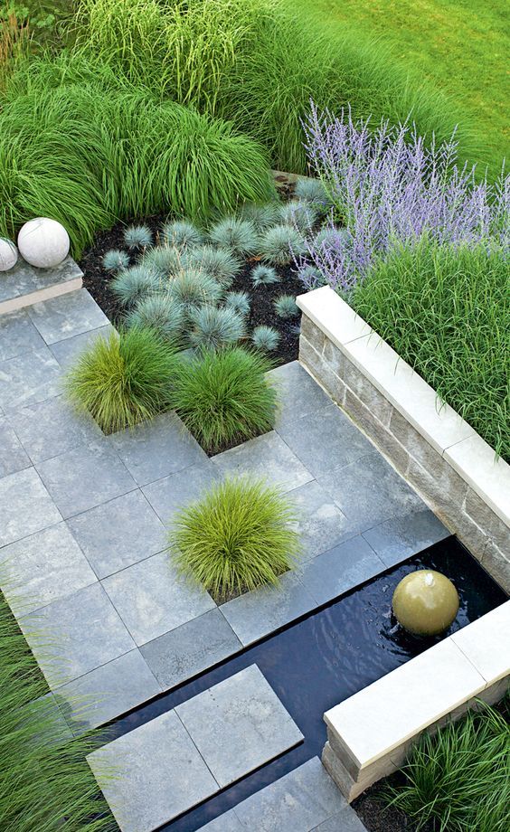 a modern outdoor space with lots of grasses and plants, with stone pavemets and a small water feature with a large rock