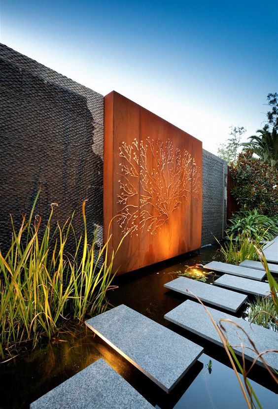 an ultra-modern outdoor space with a jaw-dropping orange accent wall with art, a large lit up water feature with stone pavements and water plants