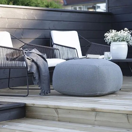 a Scandinavian deck with black metal chairs, white upholstery, grey blankets and a large pouf, some potted blooms