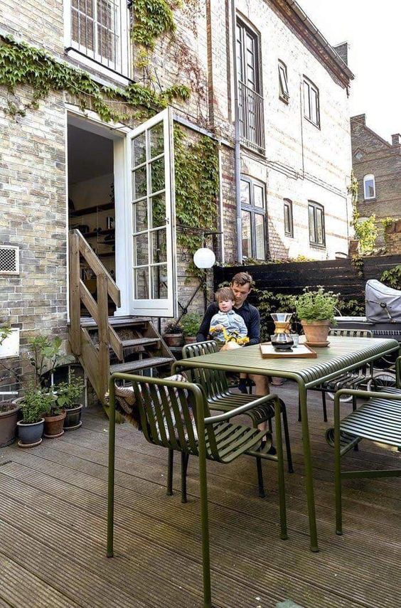 a Scandinavian patio with a wooden deck, grene metal furniture, potted plants and greenery and some pillows