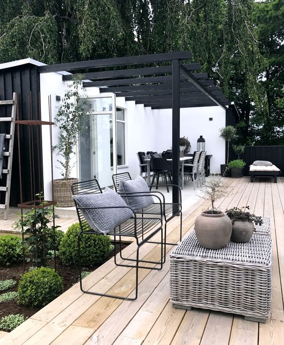 a Scandinavian terrace with a wooden deck, a woven coffee table, black metal chairs, a corner bench and greenery around