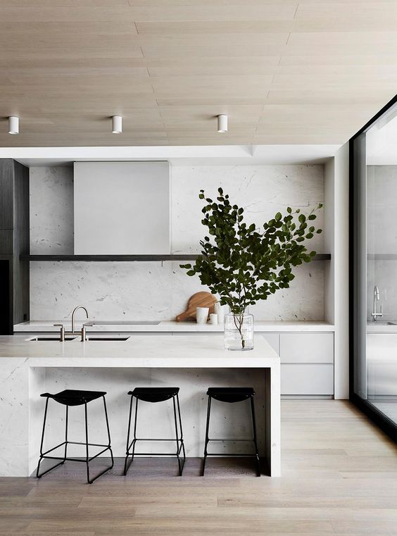 a beautiful minimalist kitchen with no hardware neutral cabinets, a white stone backsplash and countertops, a kitchen island and a glazed wall