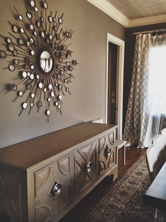 a beautiful taupe dining room with a gorgeous sunburst mirror, a credenza, a dining table and chairs, printed curtains and a ceiling