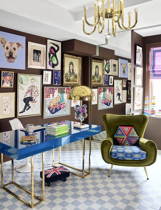 a bright and catchy home office with brown walls, a bold blue desk, a green chair with pillows, a colorful gallery wall and a large mirror