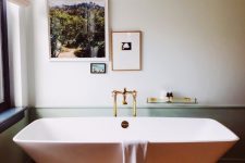a chic and peaceful bathroom with green paneling, a catchily shaped bathtub, gold fixtures and a mini gallery wall with photos