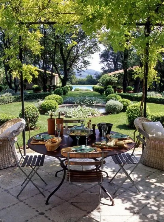 a chic and refined Provence terrace with a green lawn and greenery and trees, a round metal table and matching chairs, some woven chairs for more comfort
