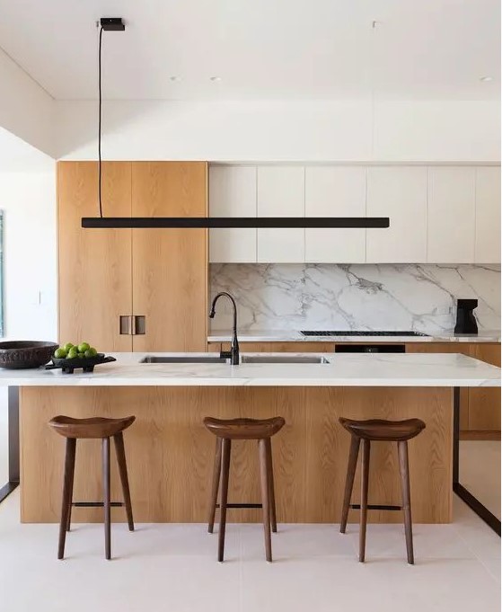 a chic contemporary kitchen with white and light stained cabinets, a large kitchen island with a white stone countertop and a backsplash, a black pendant lamp