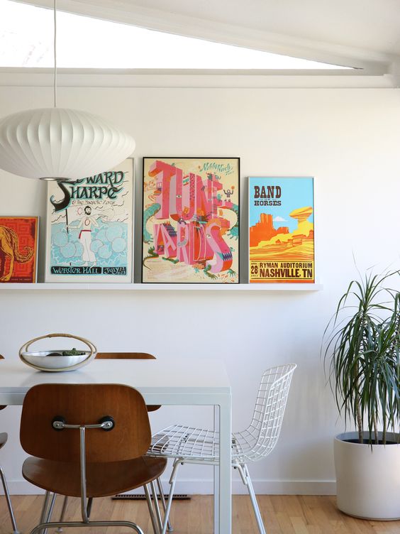 a chic mid-century modern dining room with a white table and timber and metal chairs, a ledge gallery wall with bold posters