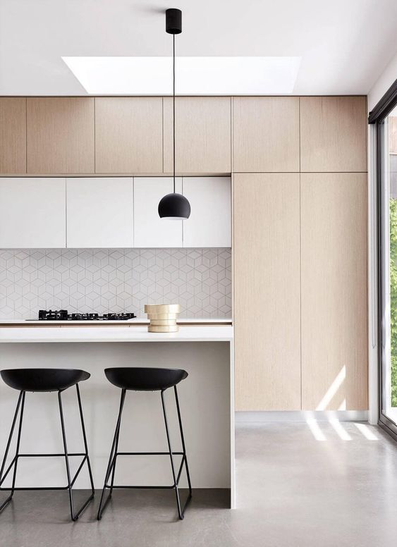 a clean line kitchen with no hardware cabinets, a white kitchen island, a geo backsplash and black stools and lamps