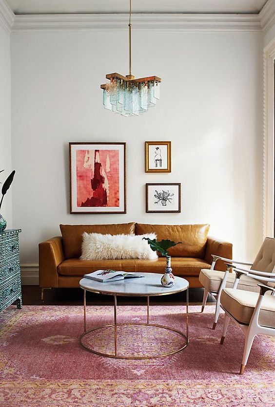 a colorful living room with a rust-colored leather sofa, beige chairs, a round table, a pink rug and a blue inlay dresser, a whimsical mini gallery wall