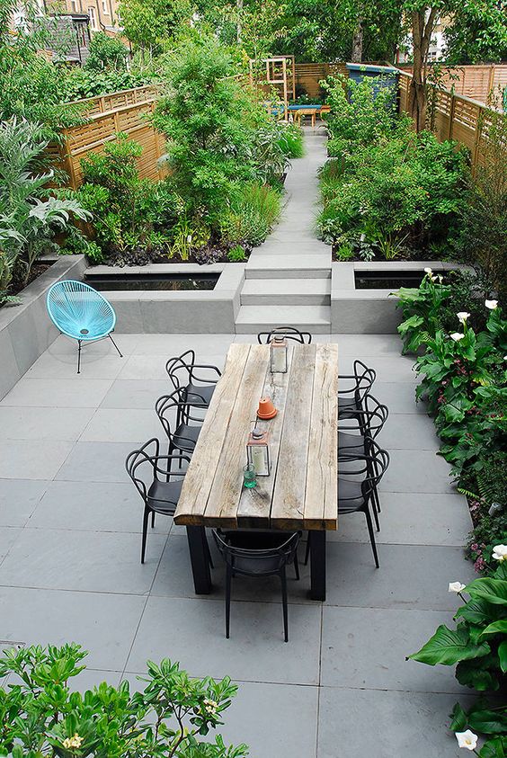 a contemporary garden with lots of greenery and blooms, clad with concrete tiles, a wooden table and black chairs