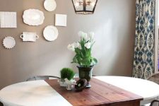 a cozy rustic dining room with taupe walls, a gallery wall of plates, an oval table, metal and woven chairs and a cool lantern