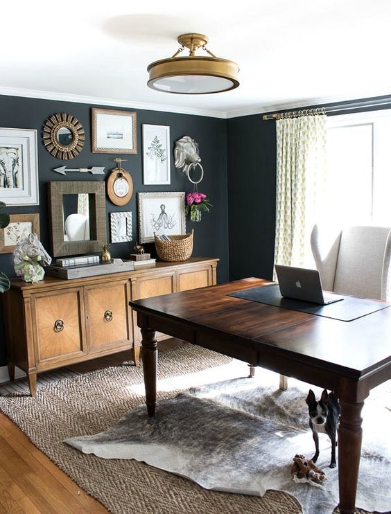 a lovely vintage home office wiht black walls, a vintage dark-stained desk, a creamy chair, a credenza and a rustic gallery wall
