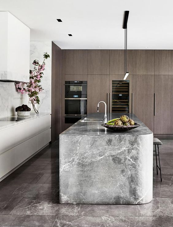 a luxurious kitchen with no hardware cabinets, a white stone backsplash and a grey stone slab island, dark-stained storage cabinets