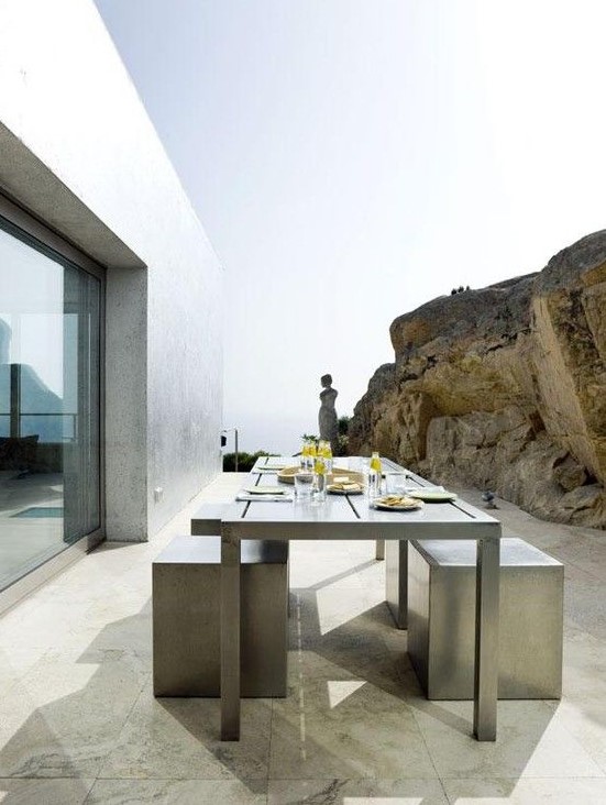 a minimalist terrace next to the rock, with a metal dining table and concrete stools plus a sea view is a cool space