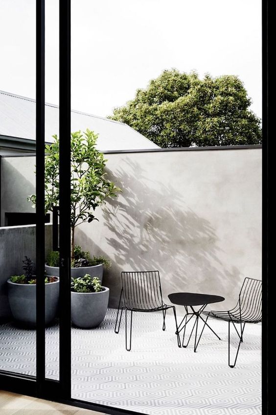 a minimalist terrace with a tall fence, a black metal dining set, potted plants in large pots and a mosaic tile floor