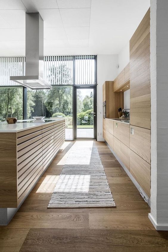 a modenr light-stained kitchen with no hardware cabinets, a wood slat kitchen island, a white backsplash and a large hood